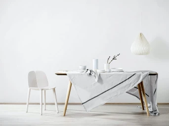 Minimalist dining table set with white tableware and a draped grey linen tablecloth beside a single white chair.