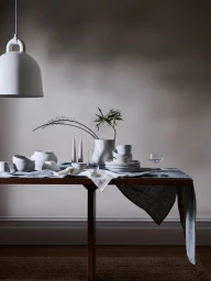 Still life table styling with ceramic tableware, plates, bowls, a vase with foliage, and a pendant lamp by Lara Hutton