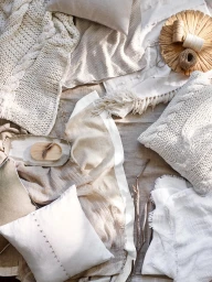 Overhead flat lay of textured neutral blankets, cable knit pillows, and wooden styling props on linen.