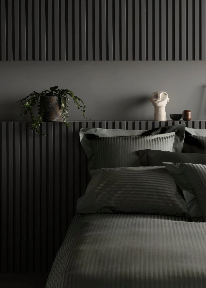 A neatly made bed with dark grey striped bedding against a wall with vertical slat panels and a potted trailing plant.