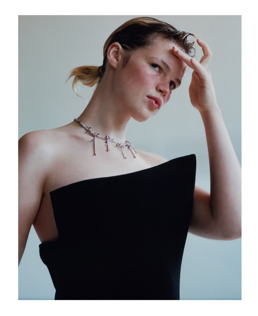 Indoor portrait of a young adult woman with fair skin wearing a black strapless top and necklace, looking slightly upward with hand raised.