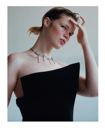 Indoor portrait of a young adult woman with fair skin wearing a black strapless top and necklace, looking slightly upward with hand raised.
