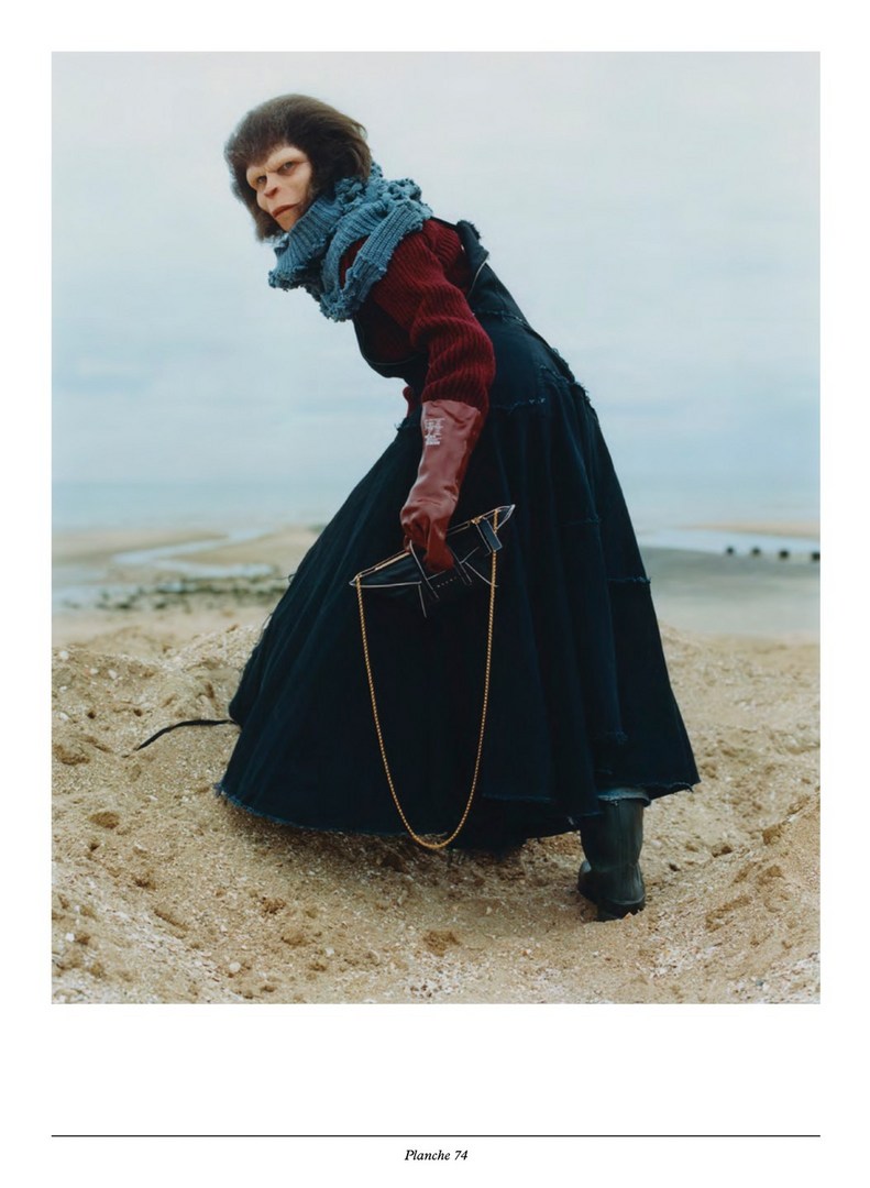 Full shot of a human in a monkey mask, a red top and black dress holding a broken mirror on a sandy beach with an overcast sky.