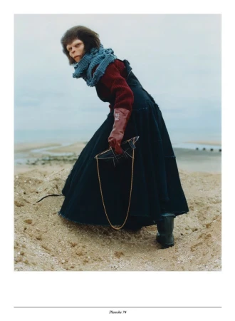 Full shot of a human in a monkey mask, a red top and black dress holding a broken mirror on a sandy beach with an overcast sky.