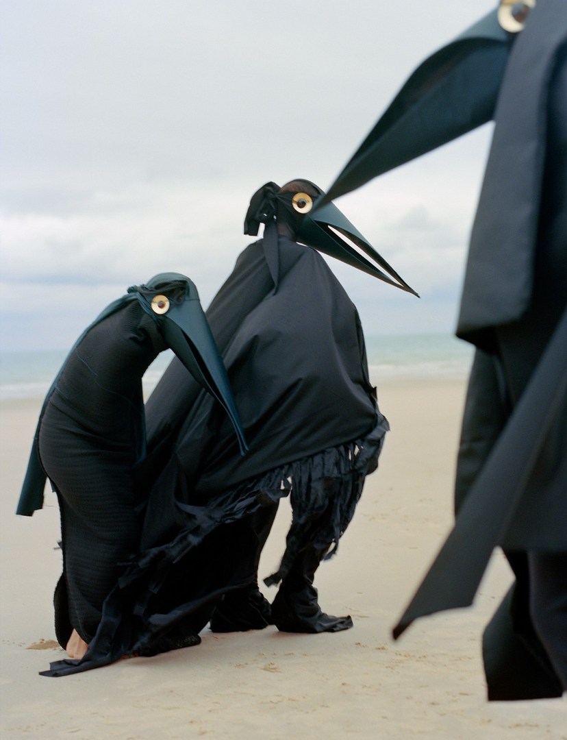 Three people in black, bird-like costumes stand on a sandy beach under an overcast sky, artfully staged for a unique fashion shoot.