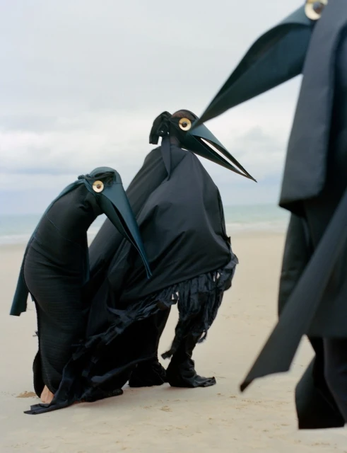 Three people in black, bird-like costumes stand on a sandy beach under an overcast sky, artfully staged for a unique fashion shoot.