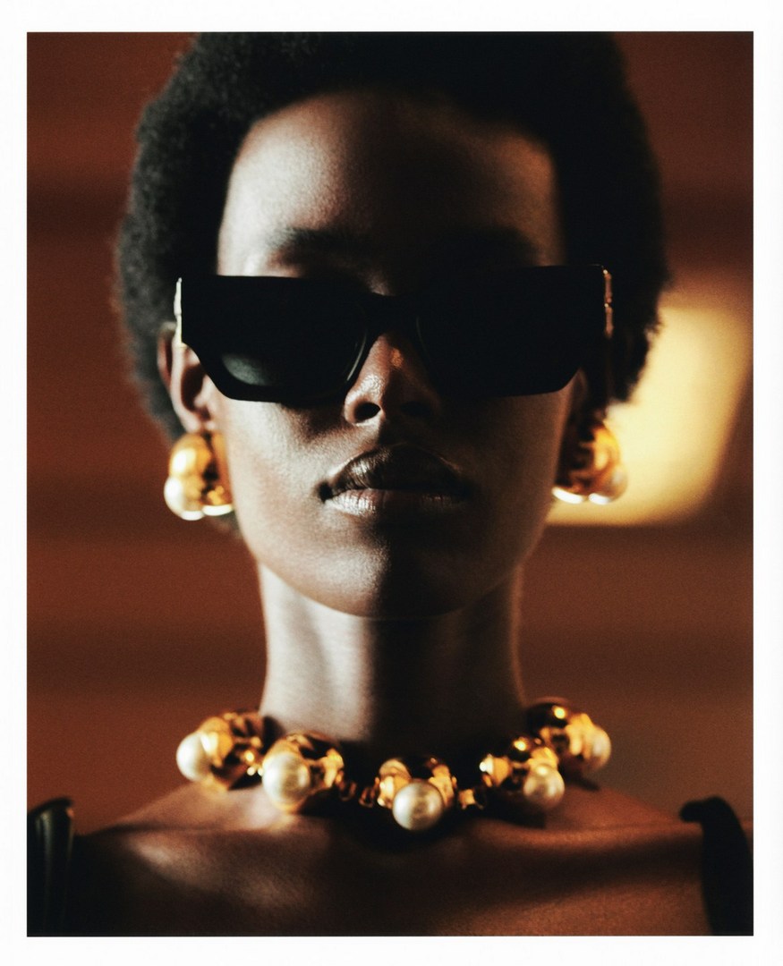 Close-up studio shot of a dark-skinned adult woman wearing large black sunglasses, a statement necklace and sculptural gold earrings.