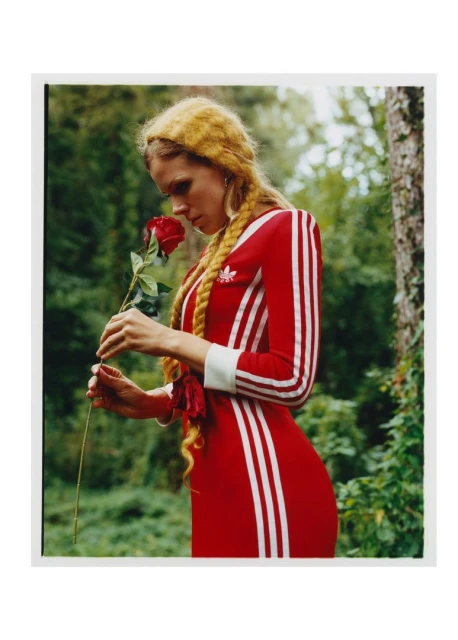 A woman in a red dress holds a red rose in a forest. The model has blonde braids and is posed in a natural outdoor setting.