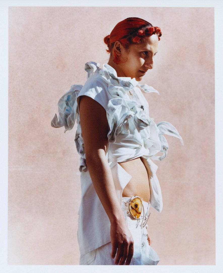 Portrait of a red-haired adult female model in an open white top with decorative ruffles, a light skirt, and light pink backdrop.