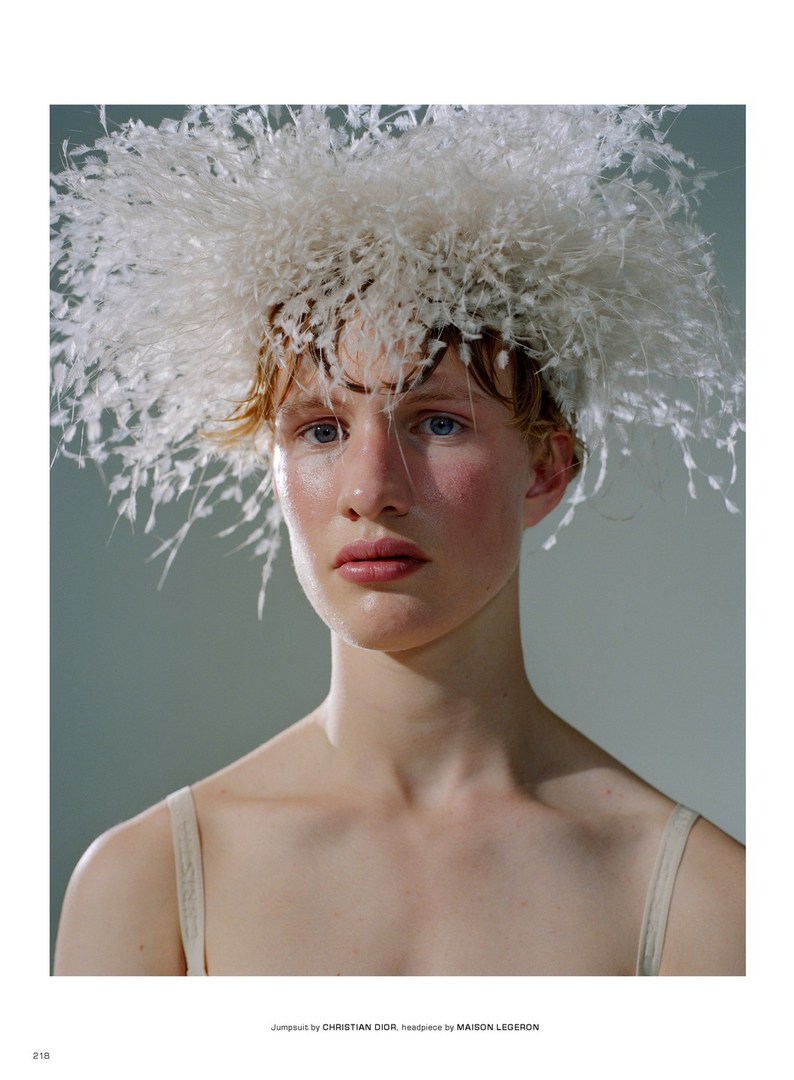 Close-up studio portrait of a woman with fair skin, wearing an elaborate feathered headpiece and a strappy top, against a gray backdrop.