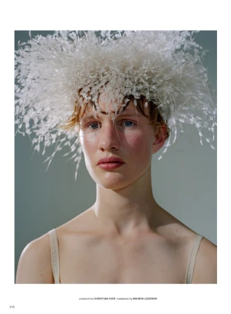 Close-up studio portrait of a woman with fair skin, wearing an elaborate feathered headpiece and a strappy top, against a gray backdrop.