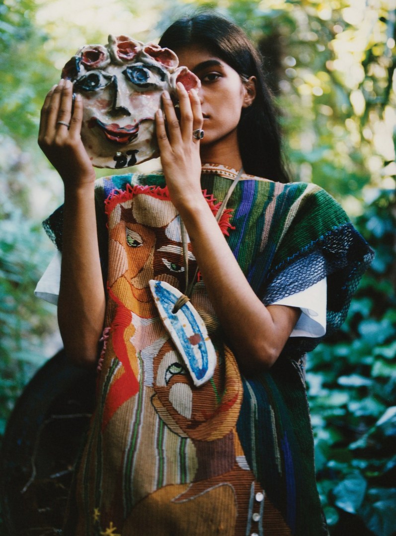 Young woman holds a ceramic face mask in front of her face while wearing a colorful patterned poncho in an outdoor setting.