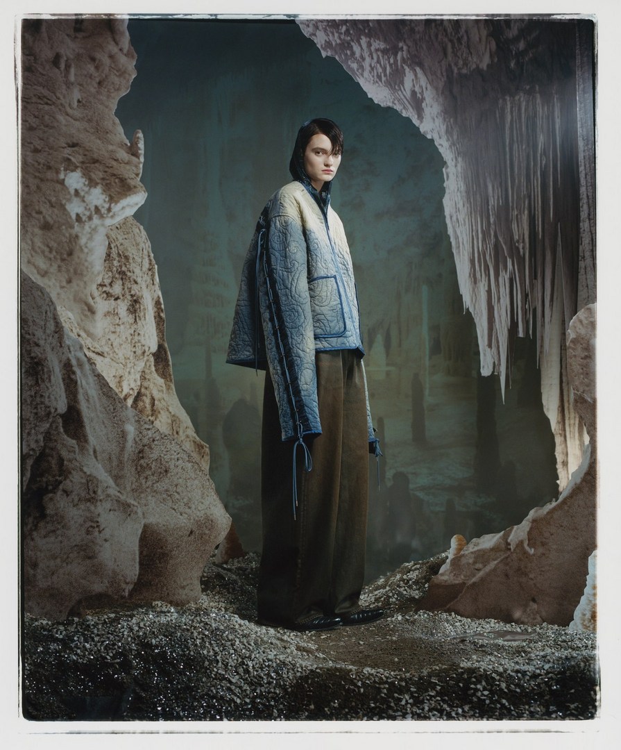 A fashion shot features a model posing in a quilted jacket and wide-leg trousers, set against the backdrop of a dimly lit cave interior.