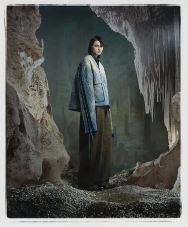 A fashion shot features a model posing in a quilted jacket and wide-leg trousers, set against the backdrop of a dimly lit cave interior.
