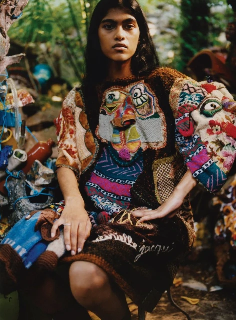 Woman wearing a colorful patterned dress seated against a cluttered outdoor backdrop with dense foliage, creating an artistic, eclectic portrait.