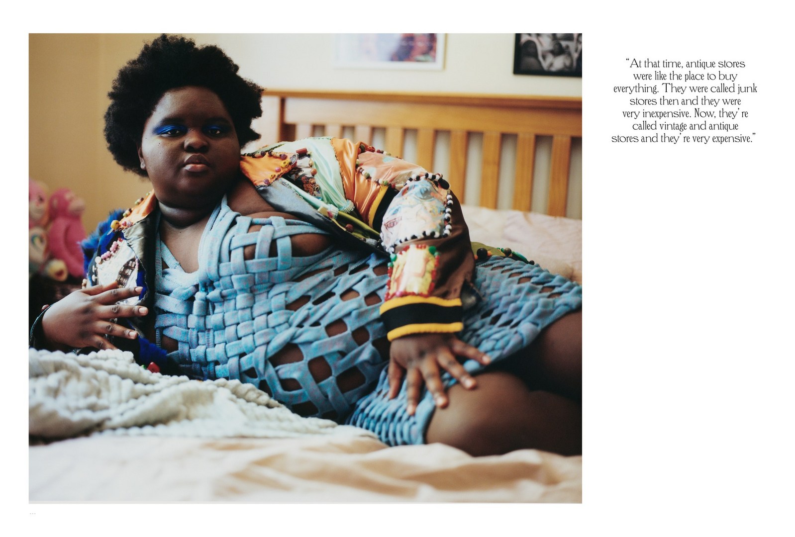 Woman with dark hair and blue eyeshadow lounges on bed with blue woven-style top and patterned jacket in a bedroom setting.