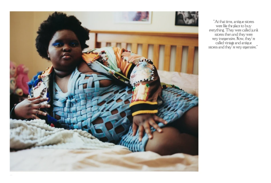 Woman with dark hair and blue eyeshadow lounges on bed with blue woven-style top and patterned jacket in a bedroom setting.