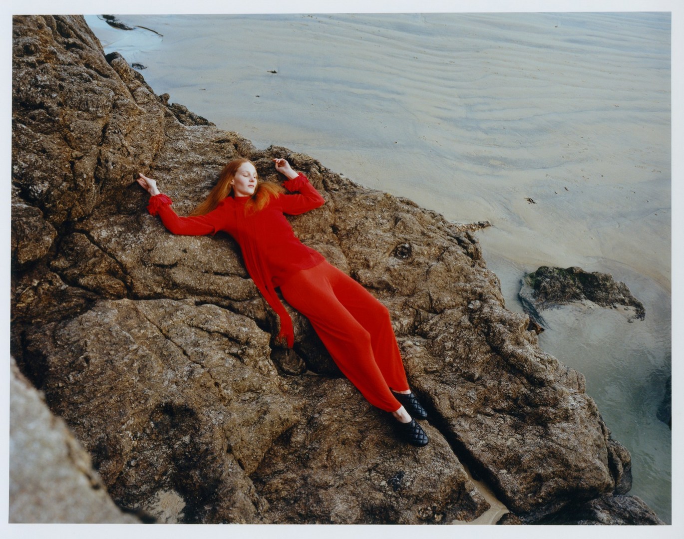 Woman in a vibrant red outfit poses on rugged coastal rocks beside a sandy beach and blue water, creating a striking contrast and scene.