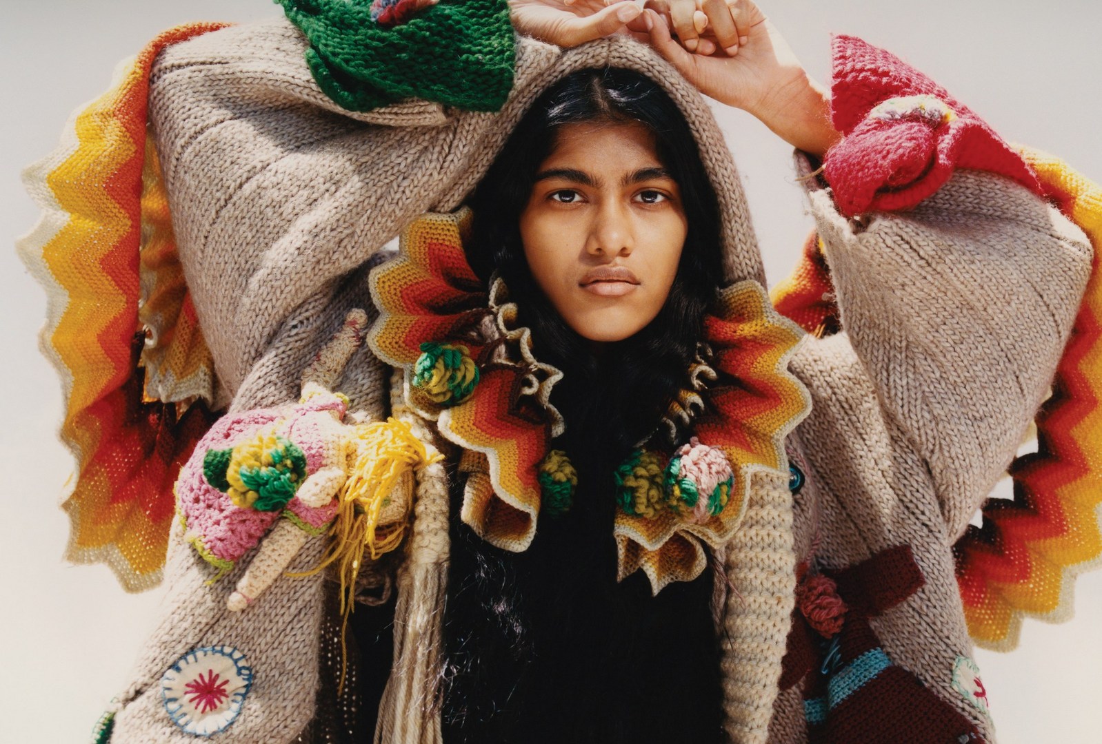 Portrait of a person with dark hair wearing a knitted, hooded garment with elaborate textile embellishments and a ruffled collar.