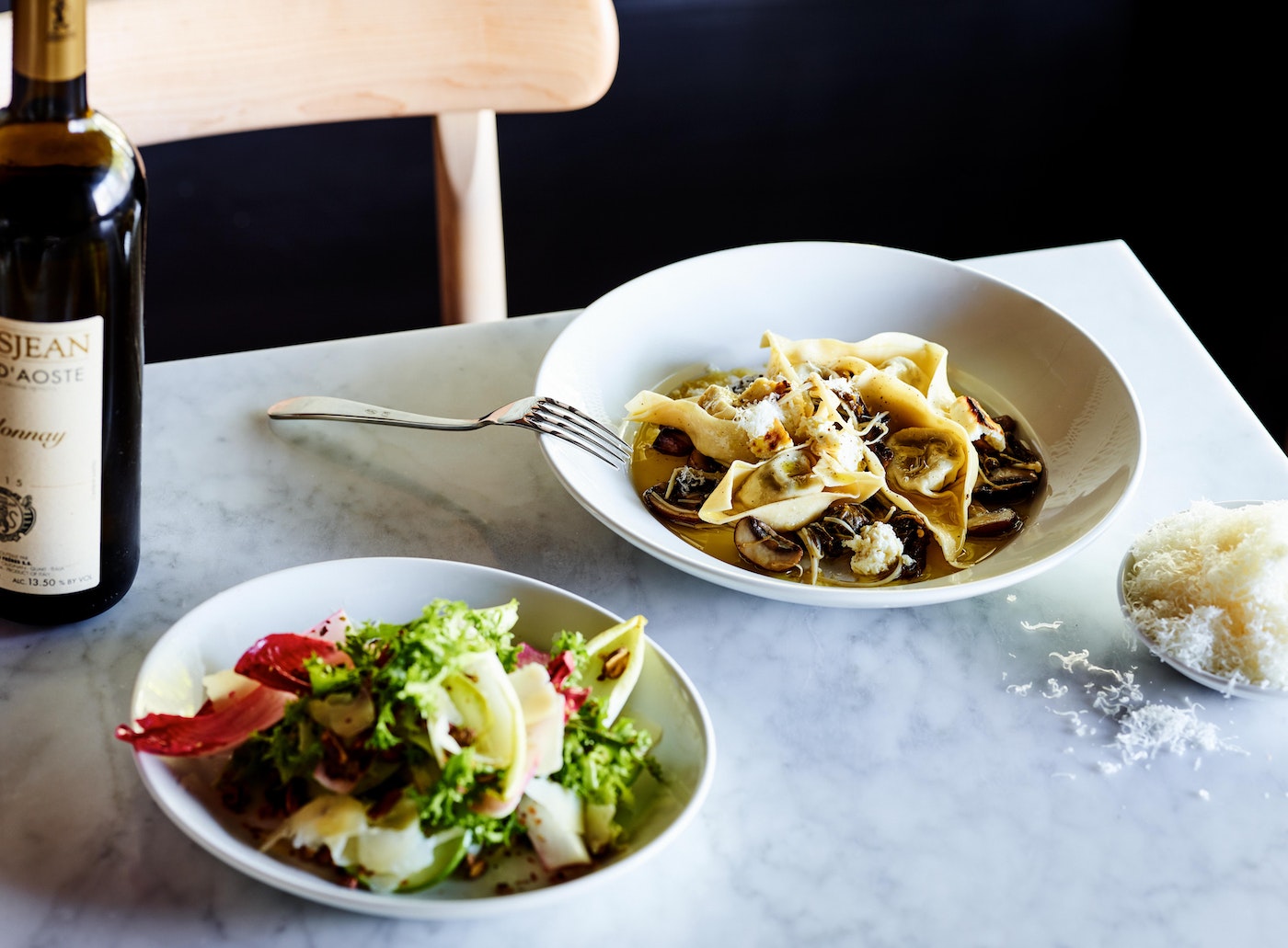 Overhead shot of white plates holding tortelloni pasta, salad, and grated parmesan cheese next to a wine bottle on a marble table.