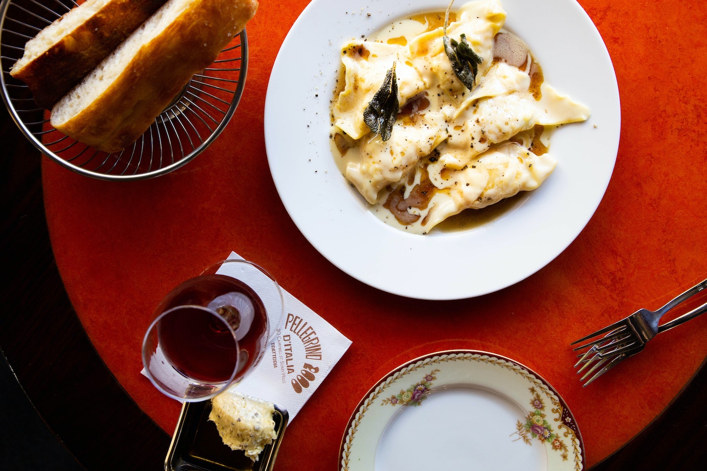 Overhead shot of tortellini pasta with sauce and sage on a white plate, alongside bread, wine, and other tableware, on a red table.