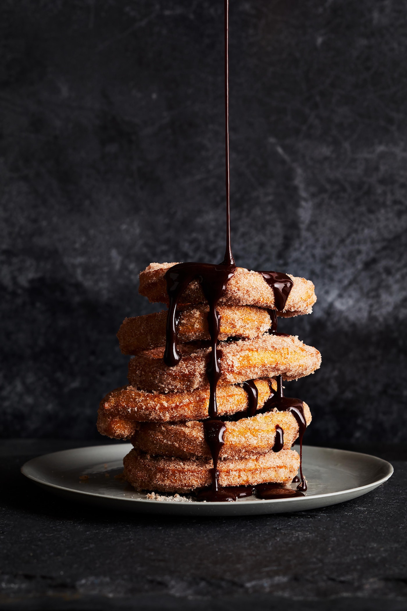 Tower of churros topped with chocolate sauce on a grey plate against a dark background, illuminated by soft, diffused lighting.