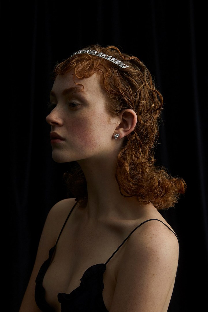 Profile view of a young woman with red curly hair wearing a jeweled headband and black top against a dark background in a studio.