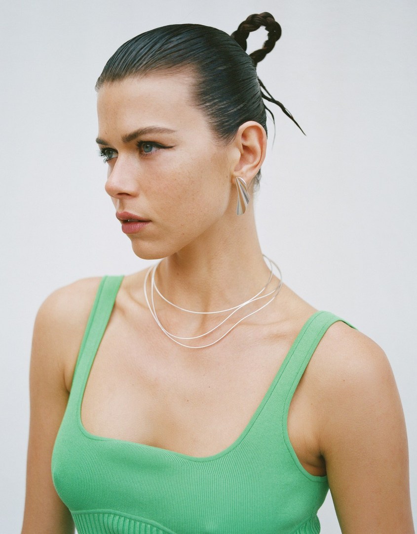 Portrait of a woman with dark hair and light eyes wearing a green top, silver necklace, and earrings against a plain white backdrop.