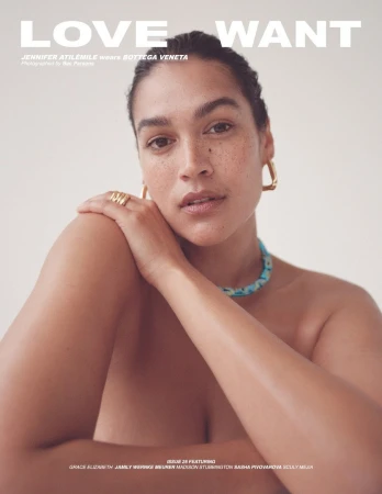 Close-up portrait of a woman with freckles and dark hair styled back, wearing gold hoop earrings and a patterned necklace against a pale background.