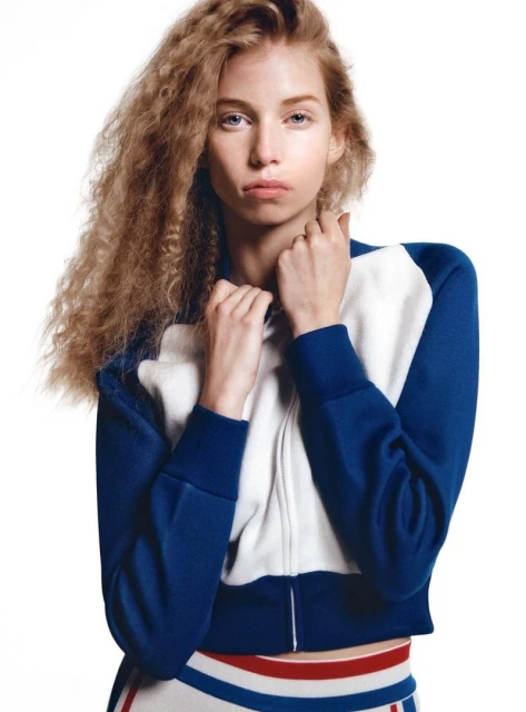 Blonde woman in a sporty jacket and skirt poses in a brightly lit photo studio, showing a casual yet fashionable look for sportswear.
