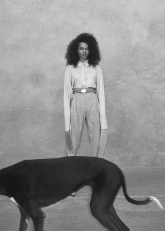 Portrait of a woman with curly hair wearing a long-sleeved button-down shirt and high-waisted trousers behind a dog
