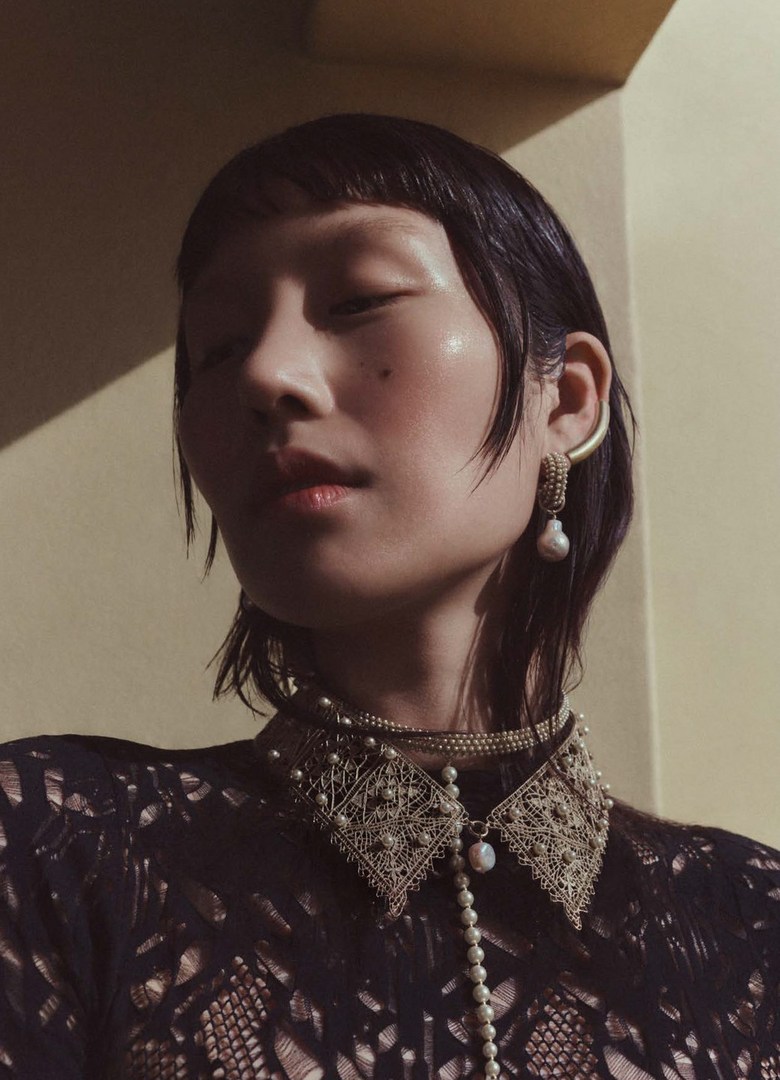 Close-up portrait of a young Asian woman with dark hair, wearing a pearl-embellished lace top and pearl jewelry in a soft-lit indoor setting.