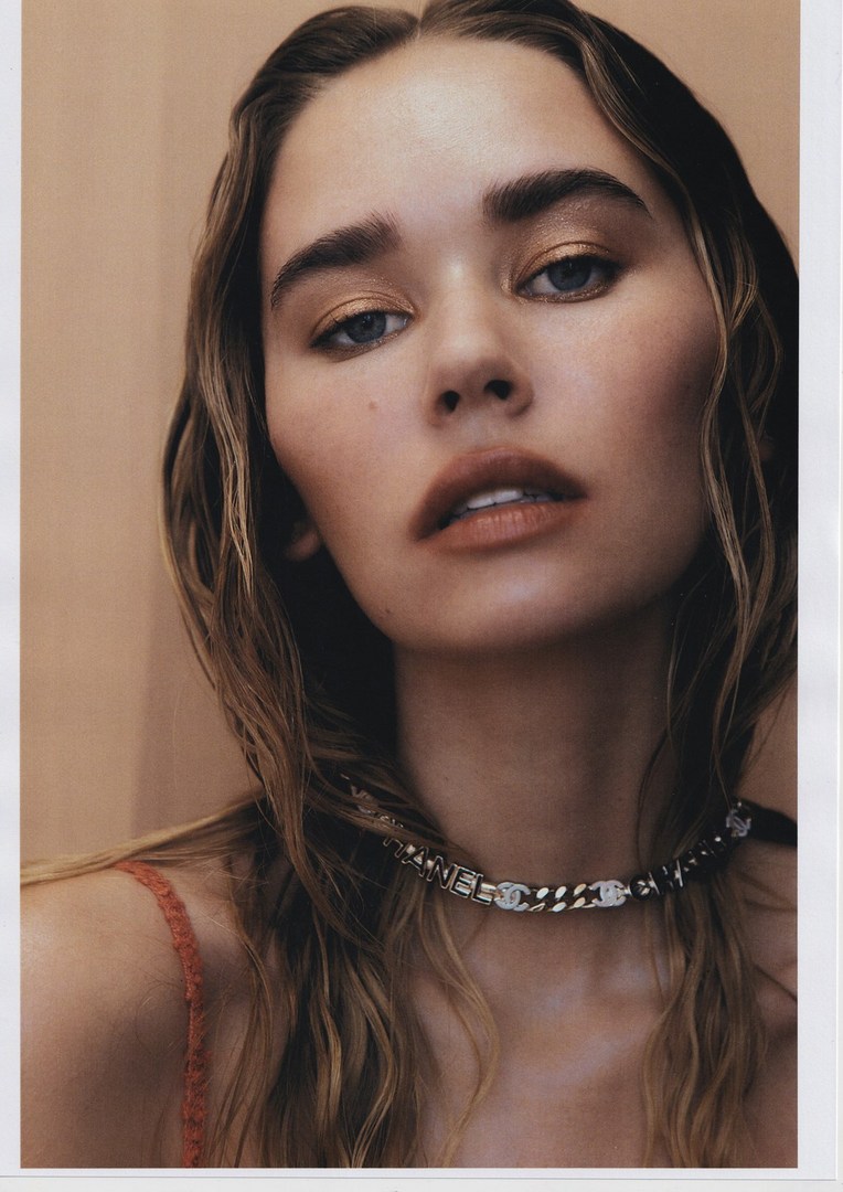 Close-up portrait of a woman with long hair and a chain necklace, soft lighting, and a peach-toned background in a beauty style.