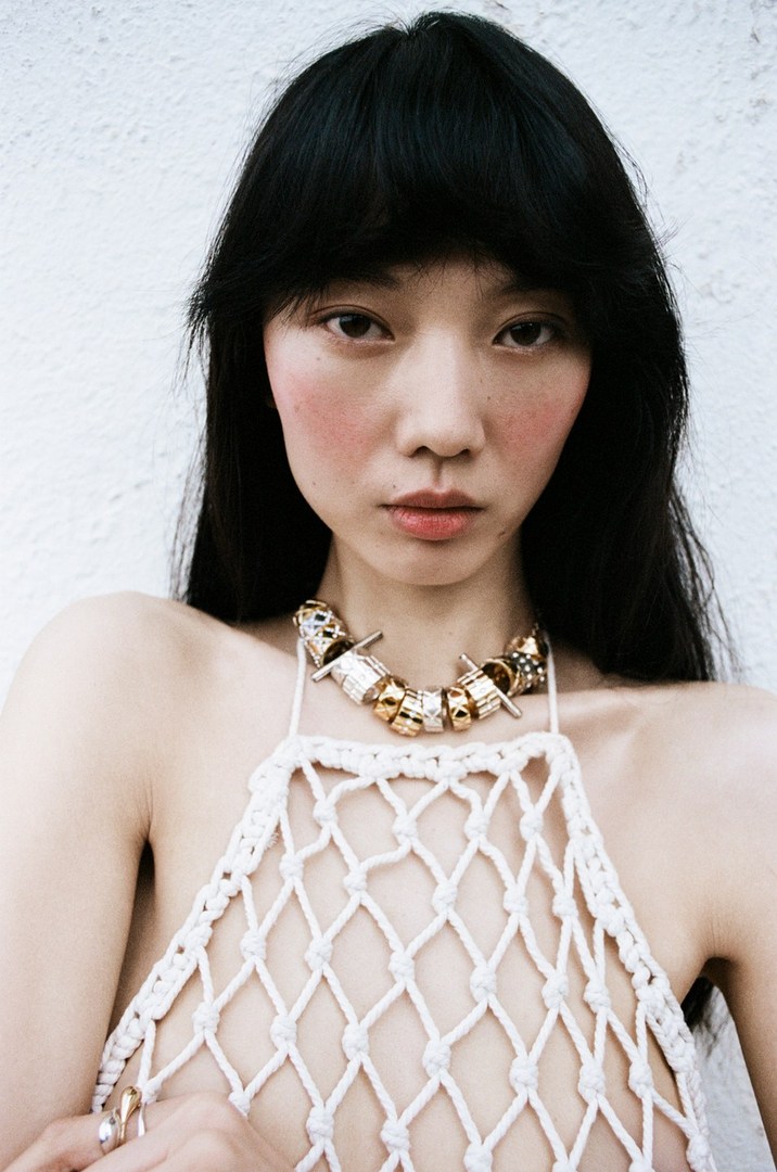 Asian woman in a white crochet top and statement necklace against a white wall, close-up portrait fashion, beauty photography.