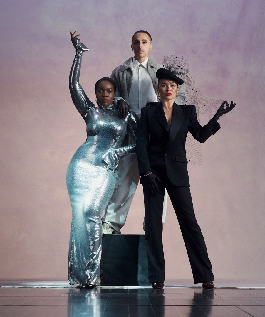 Three models pose in a studio against a purple background. An African-American woman wears a silver dress. Another woman wears a black suit and a man stands in the background.