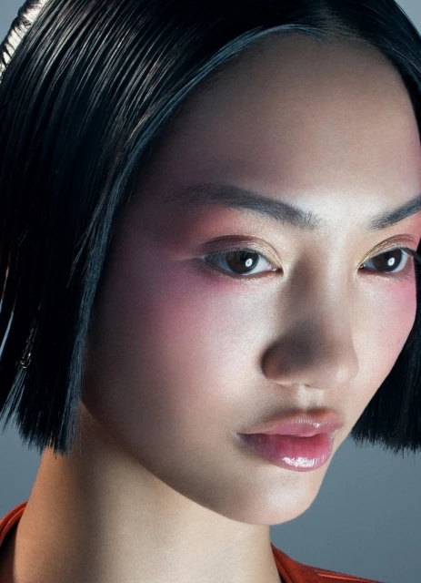 Close-up portrait of a young woman with black hair and light skin, posing against a grey background. The image showcases beauty lighting and makeup.