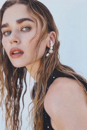 Close-up portrait of a woman with wet, wavy blonde hair wearing a pearl ear cuff against a plain white background