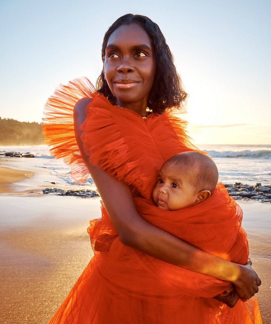 Mother holding baby in orange dress on beach at sunset. Mother looks to left, baby looks to viewer. Golden light and soft waves.