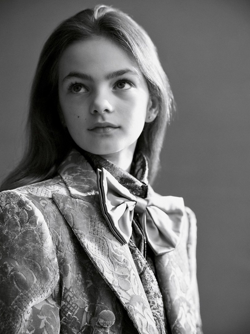 Portrait of a young girl in an ornamental jacket and bow tie, looking up and to the left in a monochrome indoor setting. 