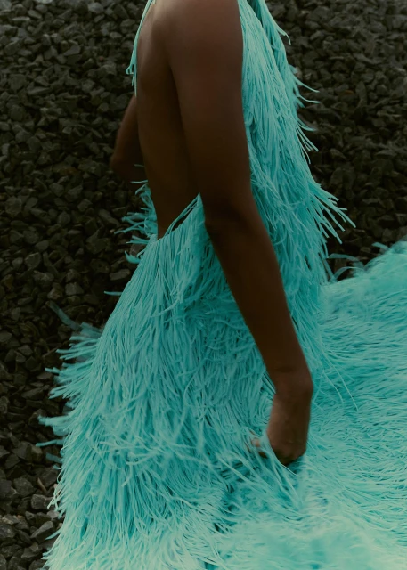 Fashion shot of a slender adult female in a flowing turquoise feathered dress, posing among jagged gray rocks. Focus on dress and texture.