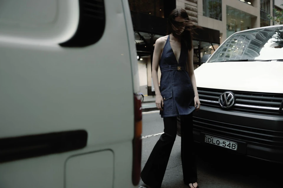 A brunette woman with long hair poses in a denim dress and black pants in front of a white van on a city street.