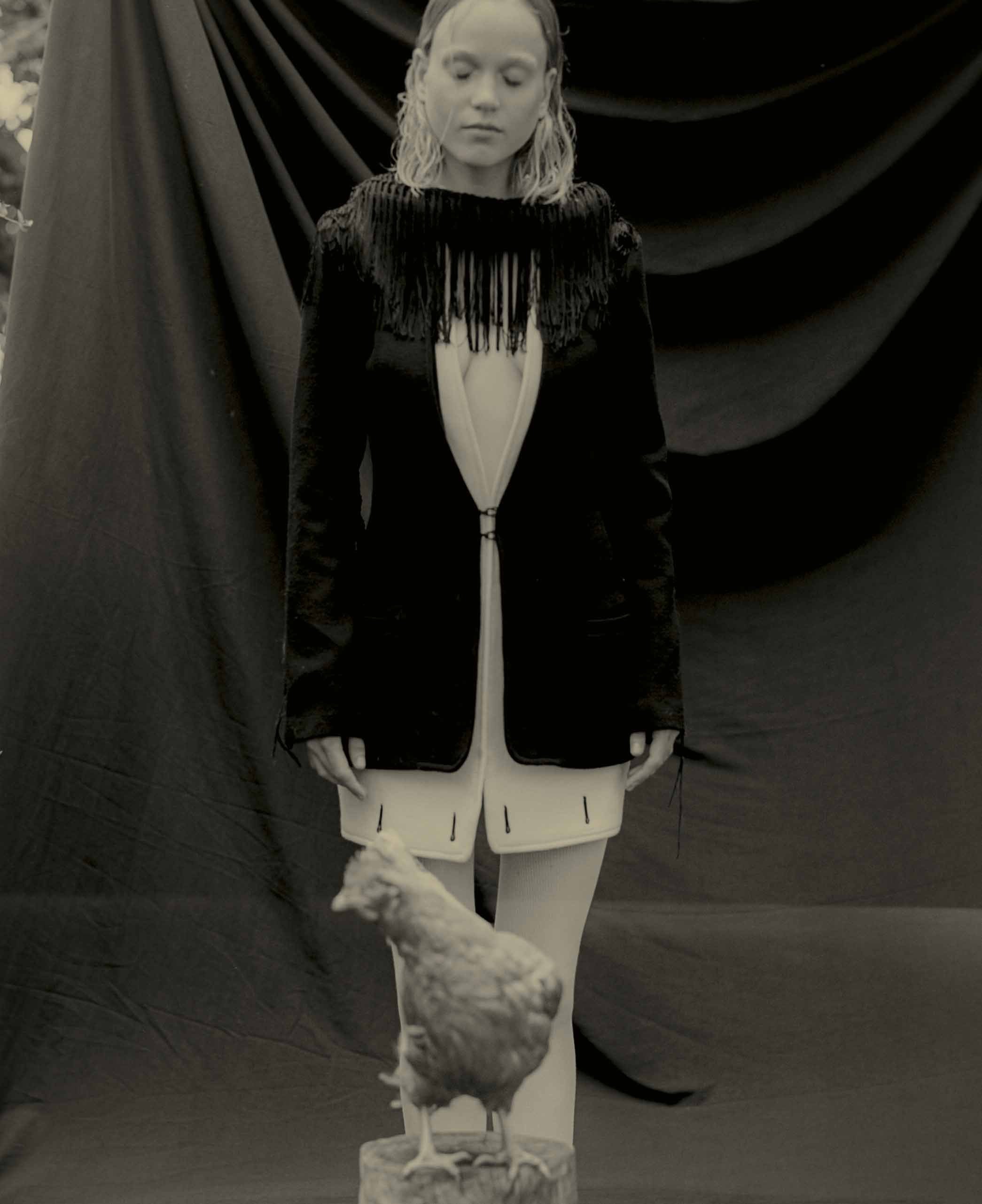 Monochrome portrait of a woman in a fringed jacket posing with a chicken standing on a tree stump in an outdoor setting.