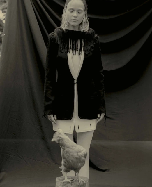 Monochrome portrait of a woman in a fringed jacket posing with a chicken standing on a tree stump in an outdoor setting.