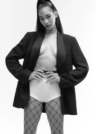 Model wearing an oversized black blazer over white shorts and fishnet tights against a bright white background