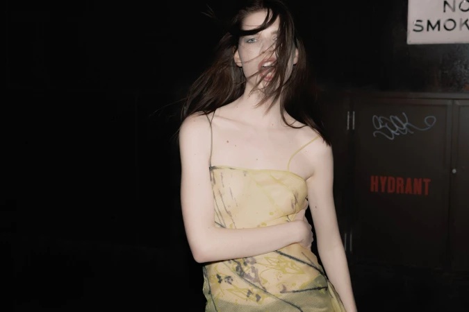 Young woman in a semi-sheer dress stands indoors against a dark background with nearby signage and graffiti, creating an edgy fashion shot.