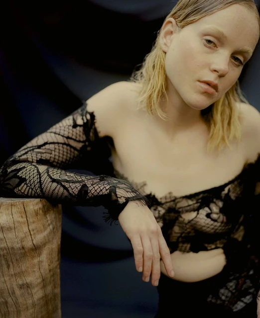 Woman in black lace top poses in front of dark backdrop next to wooden stump in studio lighting. Fashion editorial portrait.
