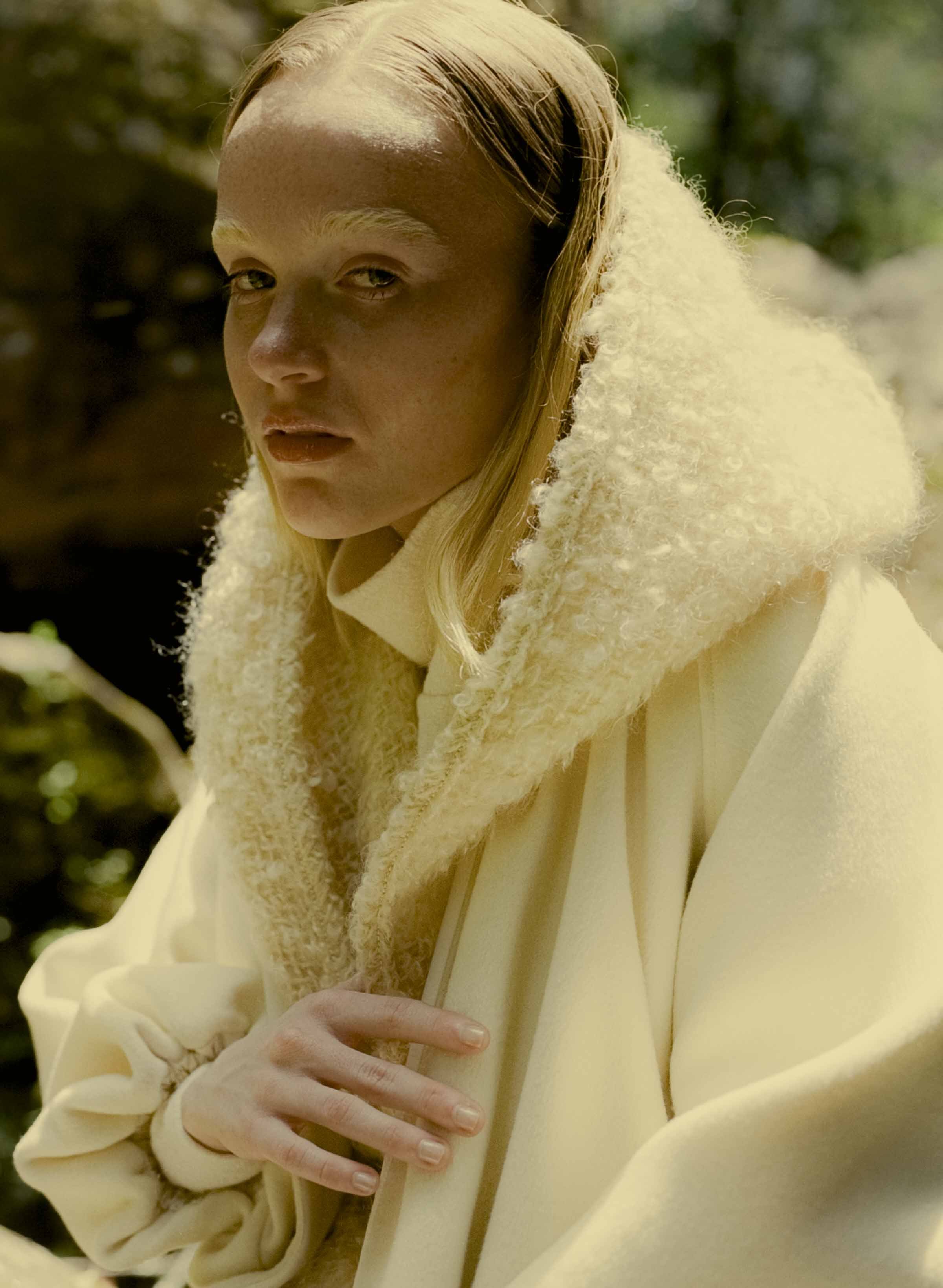 An adult female model with fair skin and blonde hair poses in nature wearing a white coat with a fluffy collar and neutral makeup.