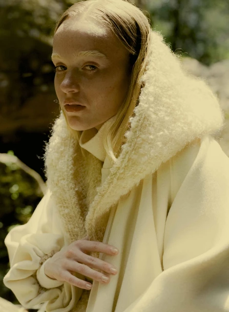 An adult female model with fair skin and blonde hair poses in nature wearing a white coat with a fluffy collar and neutral makeup.