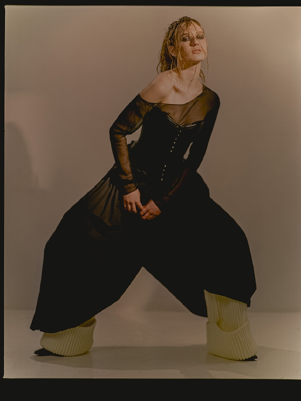 Fashionable woman poses in an off-the-shoulder long sleeve top, oversized pants, and leg warmers in a studio with neutral lighting.