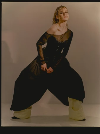 Fashionable woman poses in an off-the-shoulder long sleeve top, oversized pants, and leg warmers in a studio with neutral lighting.